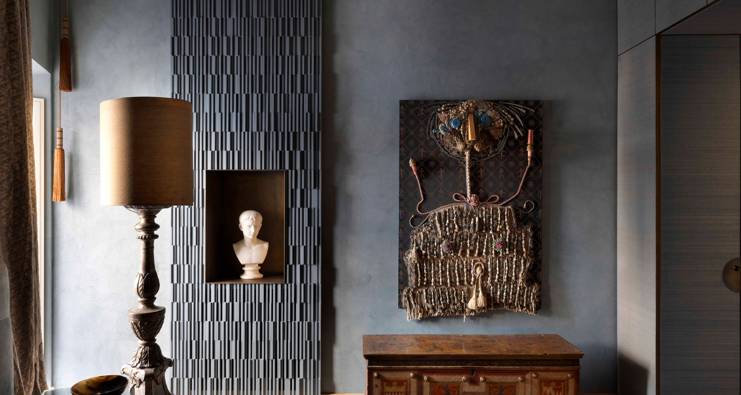 A glimpse of La Grande Bellezza, an interior project designed by Giorgia Mirabella for a historic house in Rome. Infusing opulence with contemporary styling, the image shows curtains, a lamp with a wooden base, a marble bust in a niche and an intricate woven wall hanging.